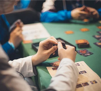 Egy csoport ember ül egy zöld asztal körül; az előtérben egy személy kezében apró fekete játékkockákat tart és rendezget.